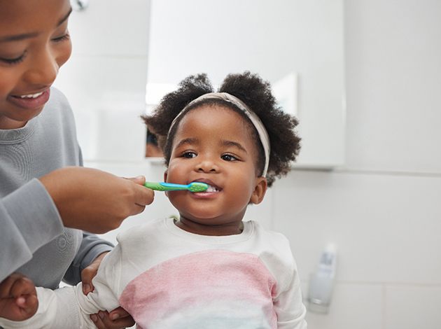 Woman in gray sweater brushing her little girl’s teeth with blue and green toothbrush